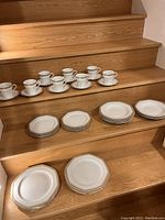 Photo of 8 cups with matching saucers arranged on stairs, along with stacks of dinner plates, dessert plates, and soup bowls showing octagonal shapes and gold rims.