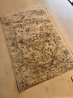 Rectangular beige patterned area rug lying on floor with tape measure along side showing size.