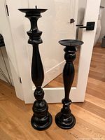 Front view of both black tall candle holders, showing sculpted detail and round base, positioned in front of glass door indoors