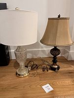 Two table lamps side by side on wooden floor against white wall, showing full height and shades.