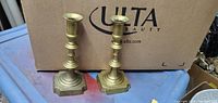 Two 8-inch brass candlesticks positioned on a blue surface with an Ulta Beauty cardboard box in the background.