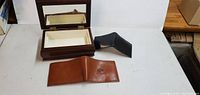Two leather wallets and one wooden jewelry box placed on a white table. The jewelry box is open showing the fabric interior.