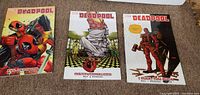 Front covers of three Marvel Deadpool graphic novels placed on carpeted floor showing colorful illustrations of Deadpool in action poses.