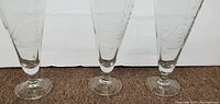Three clear pilsner glasses with etched sailboat standing in a row on a carpeted floor.