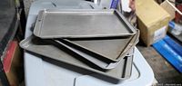 Image showing five metal baking sheets stacked on top of each other, rectangular shape with raised edges, gray metallic color.