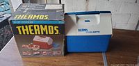 Photo of a blue and white Thermos brand plastic cooler next to its original box showing the product name and branding.