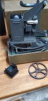 Side view of Univex PC-10 8mm movie projector inside green carrying case, showing film reel and lens cap in foreground.