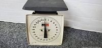 Front view of the Hanson kitchen scale showing the dial face with 0-25 lbs markings, the needle pointing to zero, and the black weighing platform on top.