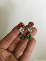 Front view of both sterling silver earrings showing red and green rhinestones with silver triangular accents, held in hand for scale.