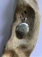 Closed view of small oval sterling silver photo locket with floral etched detail on the front, resting on a textured wooden surface.