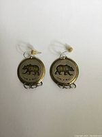 Front view of the pair of round brass and sterling silver earrings featuring bear cut-out design hung on ear wires with plastic stoppers.