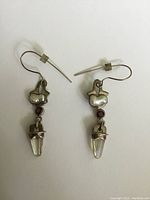 Pair of sterling silver dangle earrings against a white background, showing the pearl-shaped silver element, round garnet, and clear rock crystal pendant.