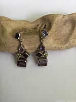 Pair of sterling silver dangle earrings displayed on a piece of driftwood showing the front view with visible colored stones in purple, green, and red hues.