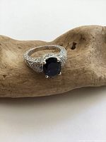 Close-up photo of the front of a sterling silver ring showing a round dark blue faux sapphire stone in a four-prong setting with pavé cubic zirconia decorated band and filigree detail.