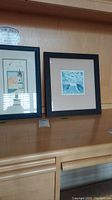 Two framed ocean art prints resting on a shelf, one showing a lighthouse and the other a coastal scene.