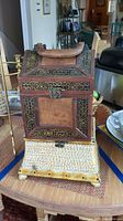 Front view of a wooden look decorative box with faux carving, nailhead trim, and top handle on a woven base.