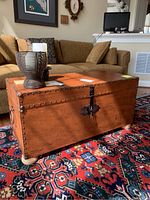 Front angled view of brown leather-like trunk coffee table, showing metal nail head trim and central locking hardware, on round wood legs, placed on multicolor patterned rug.