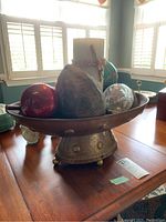 Metal vessel on pedestal filled with metal fruit, Christmas balls, and a candle viewed from front left angle.