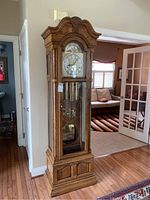 Full front view of tall wooden grandfather clock with ornate top and base, beveled glass door showing weights and pendulum.