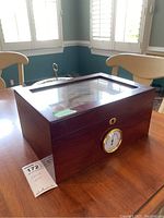 Wood humidor box on a wooden table with visible glass top lid, hygrometer on front, and a small keyhole lock.