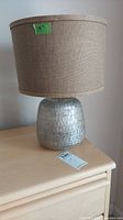 Full view of the glass based lamp with burlap type shade placed on a wooden dresser.