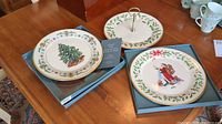 Two Lenox 1994 holiday plates displayed in blue boxes on a wooden table; one decorated with a Christmas tree and the other with Santa Claus and holly border.