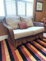 Front-side angle view of wicker loveseat with cushions and plaid pillow on multicolor striped rug