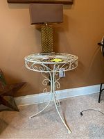 Photo of a white washed metal round table with scrollwork and a lamp with a gold textured base and brown square shade on top.