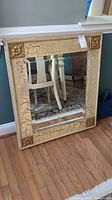 Full view of rectangular mirror with beveled edges and crackled, cream-colored frame with gold decorative corner accents resting against a wall on hardwood floor
