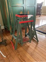 Front angle view of miniature wooden plant stand swing with green and red distressed paint on wood, showing A-frame supports and swing seat lifted off the ground on hardwood floor.