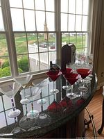 Photo showing six red martini glasses with clear stems and six clear martini glasses with decorative colored stems arranged on a granite countertop near a window.