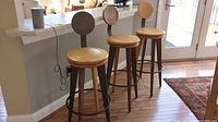 Three wooden swivel bar stools with round wooden seats, metal round backrests, metal circular footrest ring, placed near white countertop on wood floor.
