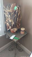 Photo showing metal corner table with ornate scrollwork, faux dried plant in planter, and small candle on table top in room corner.
