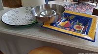 Overall view of the three serving items: stainless steel chip and dip bowl, square ceramic bowl, and hand-painted serving tray, placed on a kitchen counter.
