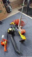 Photo of the yard tools lot showing a yellow and black blower, orange and grey string trimmer, an orange and black leaf blower nozzle, and an orange extension cord on a blue mat in a garage.