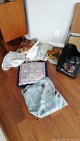 Photo showing folded fabrics, cushions, and a black tote bag on wooden floor near corner of room