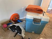 Photo showing the vintage Coleman cooler, folded orange tarp on top, pair of black vintage ice skates, weights including pink and blue dumbbells, an orange ball with netting, and a cardboard box with additional sports gear.