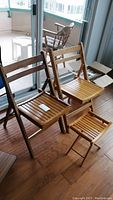Photo showing two wooden folding chairs and one folding table near a glass door. Chairs and table have slatted wood construction with visible wear and scratches.