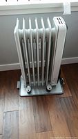 Side view of white Delonghi oil-filled radiator heater with 7 fins on metal tray base on wooden floor.