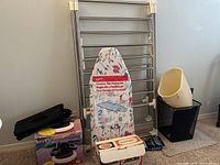 Overview of the ironing board, metal cloth rack, and two plastic dustbins on carpeted floor.