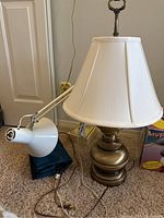 Photo showing brass table lamp with white fabric shade and white adjustable desk lamp near a door on carpet.