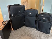 Three black Cambridge luggage pieces of various sizes with front zip pockets and wheels, standing side by side on carpet.