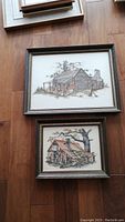 Photo showing both framed prints stacked on a floor, featuring huts and trees, in muted frames.