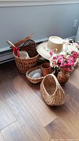 View of assorted wicker baskets, vase with artificial flowers, woven hat and trays set on wooden floor near white baseboard heater.