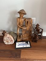 Photo showing three wooden carvings including a tall carving of a man with a conical hat and water-mill like structure, two smaller carvings of men with farming tools, two decorative ceramic flasks, and a framed display of assorted buttons