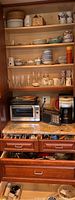 Full view of wooden cabinet with shelves containing dinnerware, glassware, woven basket and appliances below on countertop.