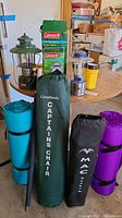 Two camping chairs in green and black carrying cases, three sleeping mats rolled in different colors, two different lanterns, a yellow container, and a black umbrella or cane on a round table in storage area.