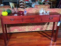 Front view of narrow wood console table with three drawers, reddish-brown finish, small glass and ceramic figurines displayed on top.