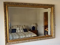 Full front view of ornate large framed wall mirror hanging on wall, showing bedroom reflection.