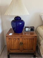 Full view of wooden nightstand with blue ceramic table lamp, pleated shade, and digital clock radio on top.